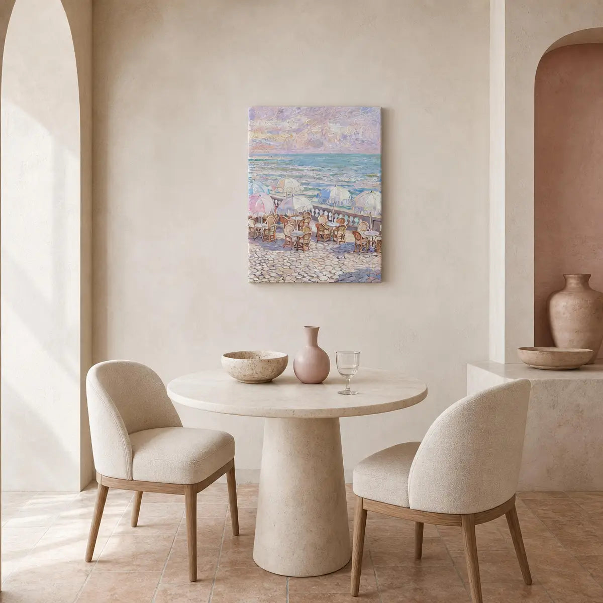 A square canvas painting of a beach scene with umbrellas and figures, hanging on a beige wall above a round stone dining table with two upholstered chairs in a softly lit interior.