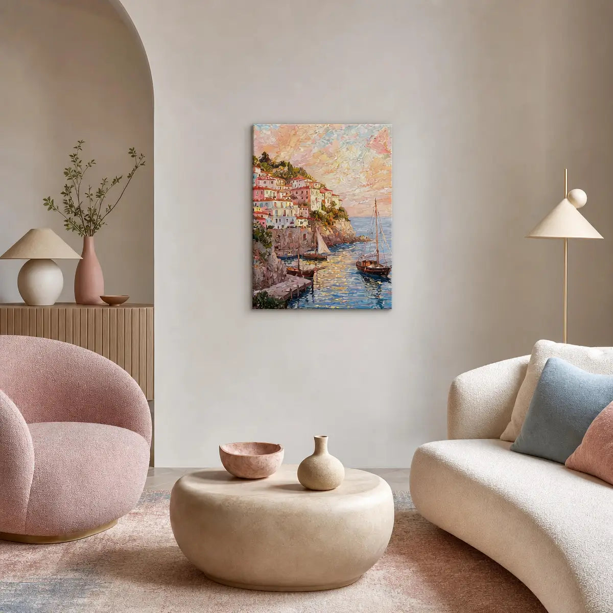 A vertical canvas art print depicting a colorful coastal village and sailboats, styled in a modern living room setting with a pink armchair, beige sofa, and neutral-toned decor.