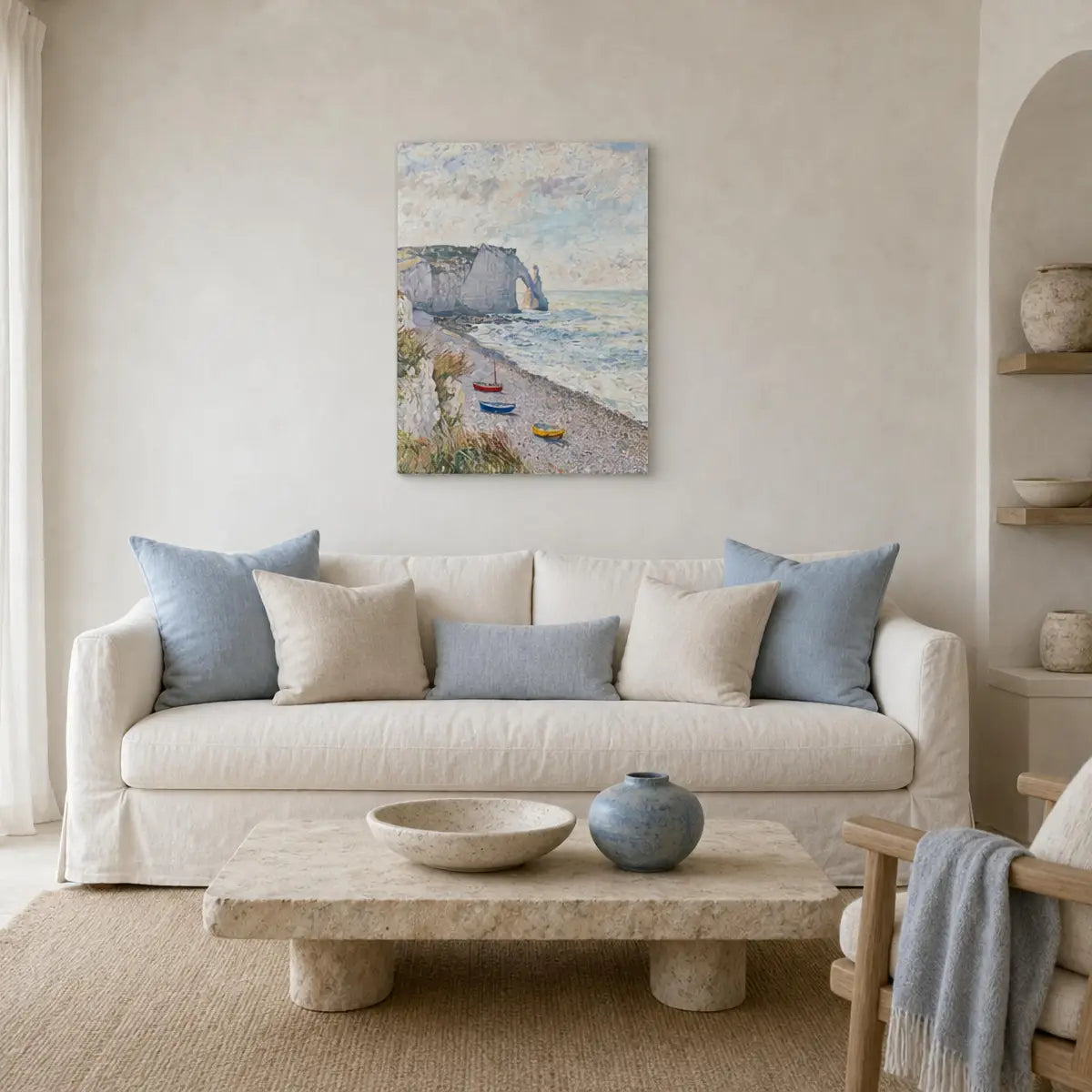 A framed coastal landscape painting hangs on a beige wall above a cream sofa with blue and beige pillows in a softly styled living room.