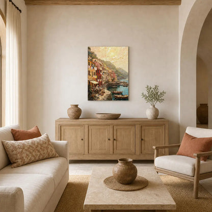 A framed canvas print of a colorful coastal village painting hangs on a beige wall in a warm, modern living room with a light wood sideboard, sofa, and armchair.