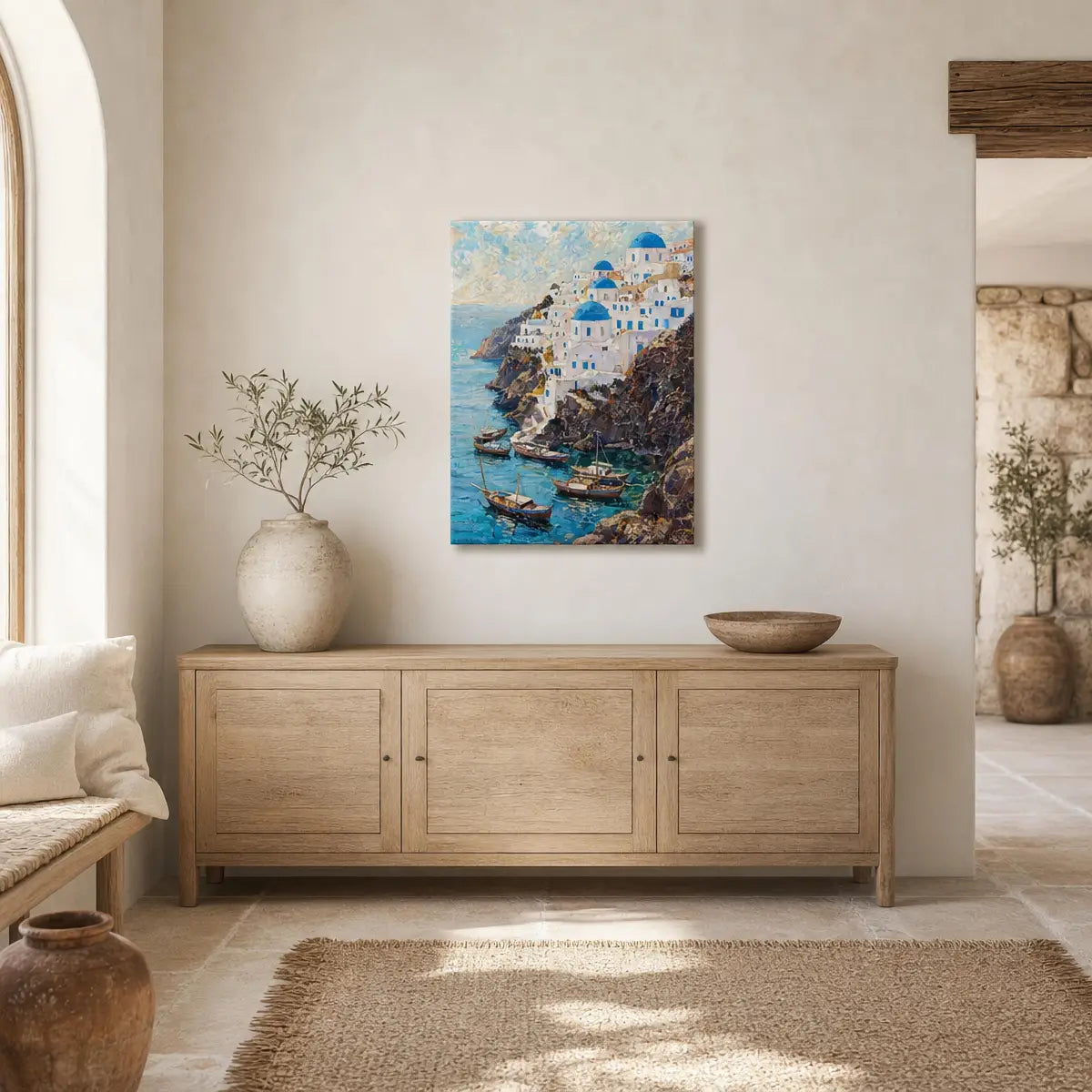 A vertical canvas print of a Santorini coastal scene with white buildings and blue domes, displayed on a beige wall above a light wood credenza in a styled living room setting.