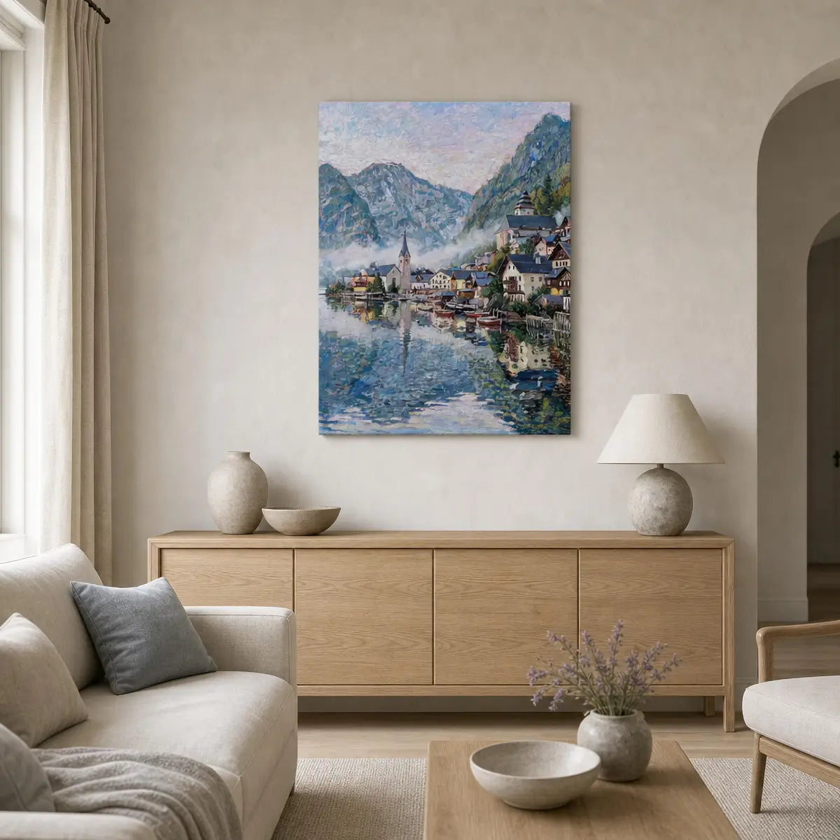 A framed impressionist-style painting of a mountain lake town hangs on a beige wall above a light wood sideboard in a styled living room with a sofa, lamp, and table.
