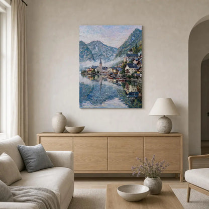 A framed impressionist-style painting of a mountain lake town hangs on a beige wall above a light wood sideboard in a styled living room with a sofa, lamp, and table.